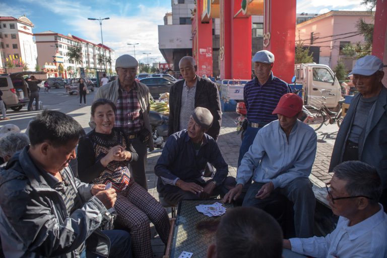 Ein Tisch und mehrere Sitzgelegenheiten, schon kann es losgehen. Viele Aktivitäten finden draußen, im öffentlichen Raum, statt. Mehrere Anwohner sitzen in einem Wohnviertel in Hulun Buir beim Karten spielen um einen Tisch, weitere Anwohnern stehen dahinter und schauen zu. Hulun Buir liegt als Teil der Mandschurei im äußerten Nordosten Chinas, unweit der Grenzregion zu Russland und der Mongolei. Hulun Buir - Innere Mongolei - China