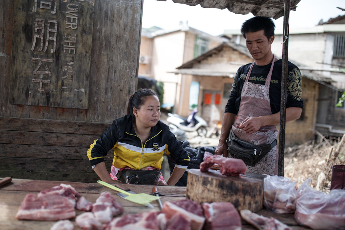 Der offene Stand der Fleischerei befindet sich direkt an einer Straße im Dorf. Die Verkäuferin wurde gerade von dem Mann beliefert. Gegen Ungeziefer liegt auf dem Tisch eine Fliegenklatsche. Yunshuiyao liegt im Süden der Provinz Fujian, eine Region in der sich viele Tolou-Lehmrundbauten befinden. Yunshuiyao - Fujian - China