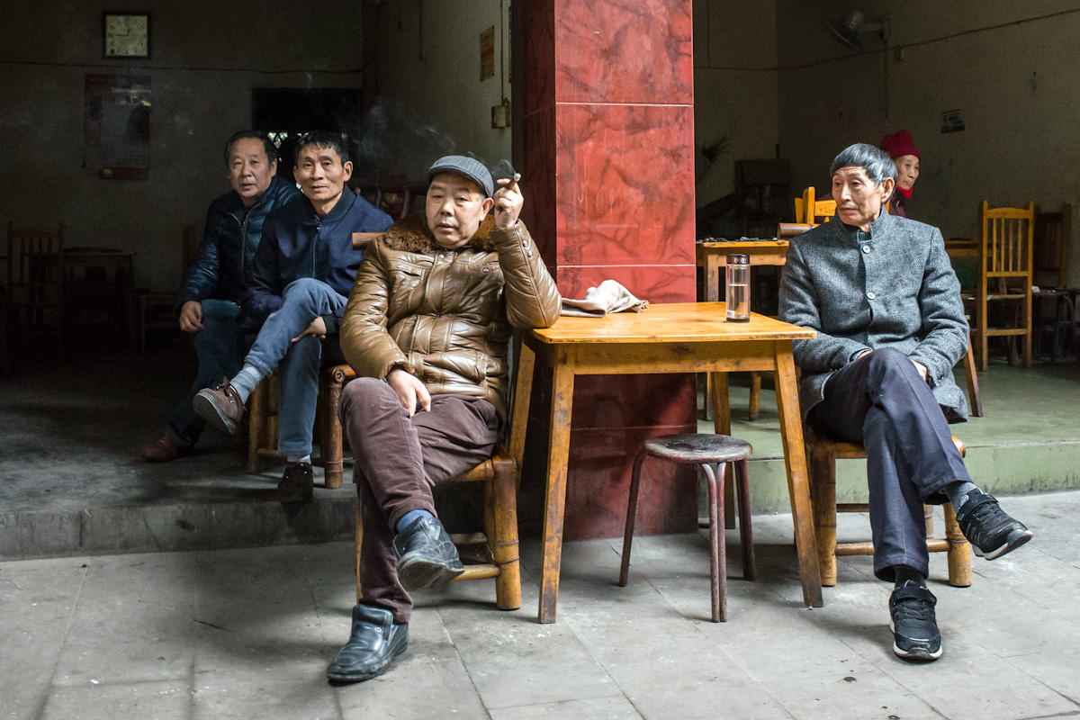 Mehrere Männer sitzen mittags in und vor einem Lokal in Qianwei und beobachten das Geschehen davor. Die Einrichtung des Lokals besteht nahezu nur aus Tischen und Stühlen und letztere aus Bambus gefertigt sind. Qianwei - Sichuan - China