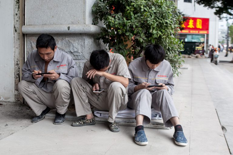 Drei Arbeiter einer Fabrik sitzen vor dem Eingang und verbringen die Mittagspause vertieft mit ihrem Smartphone. Fuzhou - Fujian - China