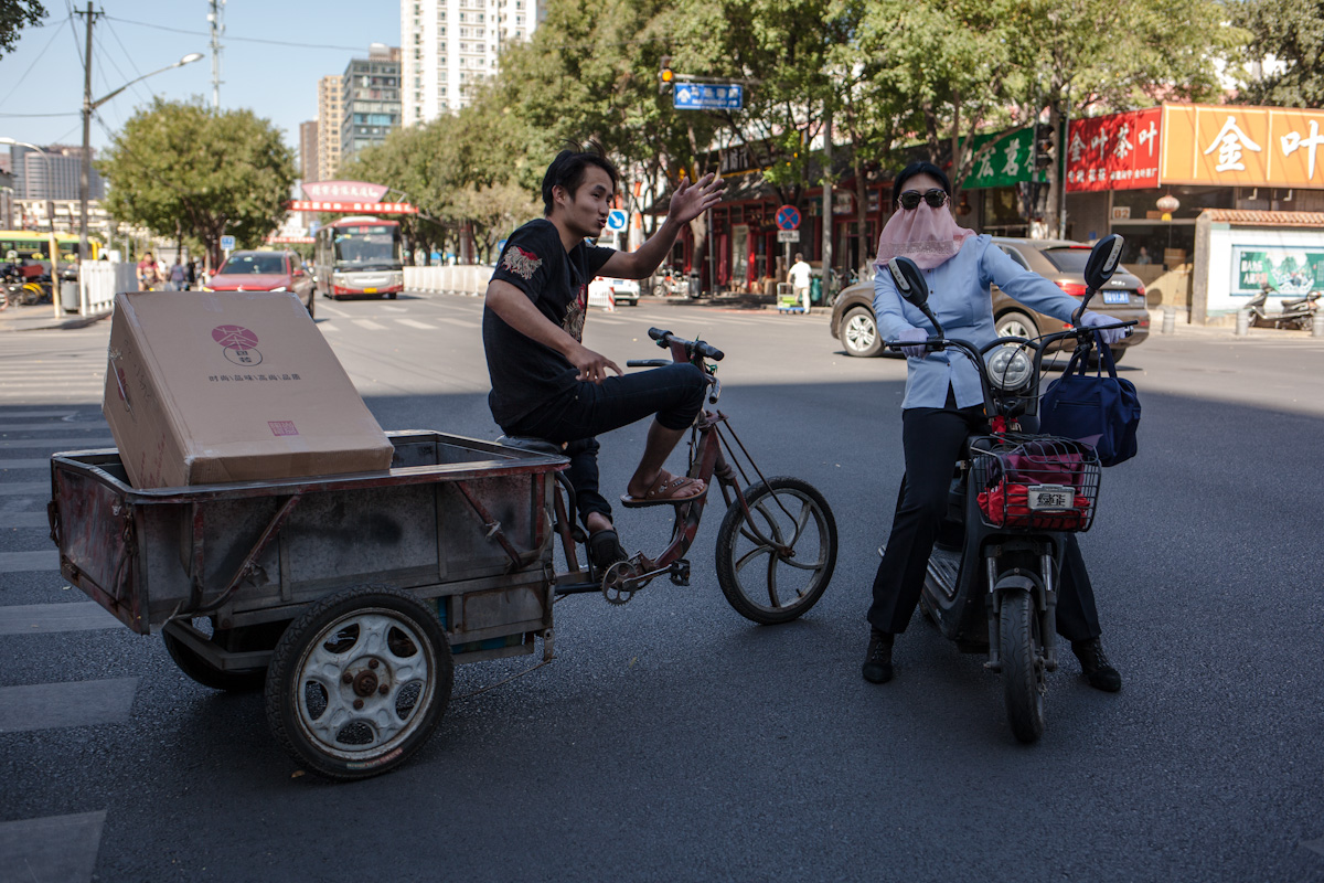 An einer Kreuzung fragt eine Frau auf einem Moped einen Mann auf einem Dreirad nach dem Weg. Mit einem Tuch vor dem Gesicht schützt sie sich vor der Hitze und Sonne. Pekig - China